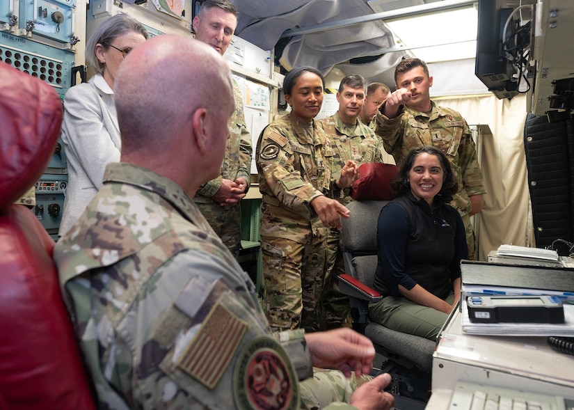 A uniformed service member speaks to a seven people while seated in a room with equipment.