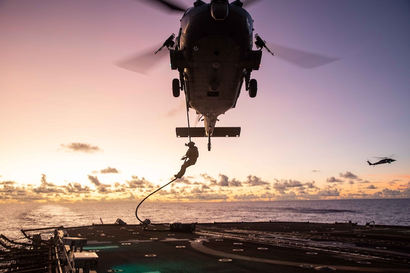 Silhouette of a sailor fast-roping from a helicopter onto a Navy ship's flight deck at twilight. Another helicopter and open water can be seen in the distance.