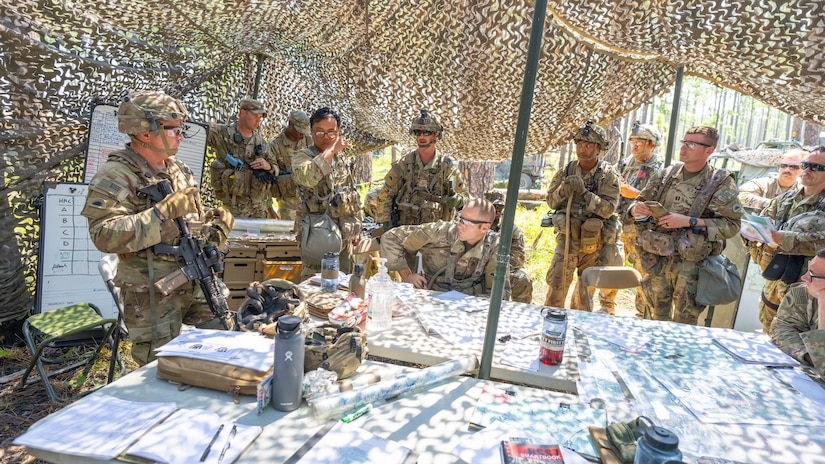 Military personnel in gear gather around a table inside of a camouflage canopy.