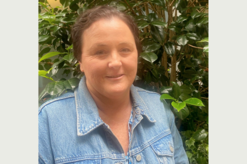 A woman smiling in a denim jacket in front of a tree.