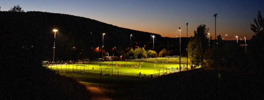 Foto av en fotballbane med gress opplyst av lyskastere. Mørk høstkveld i Norge.