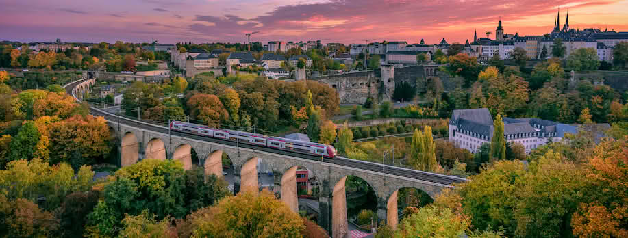 En gammel jernbanebro med søyler og buer med et tog i fart. Skyline av en by med solnedgang i bakgrunnen. Foto