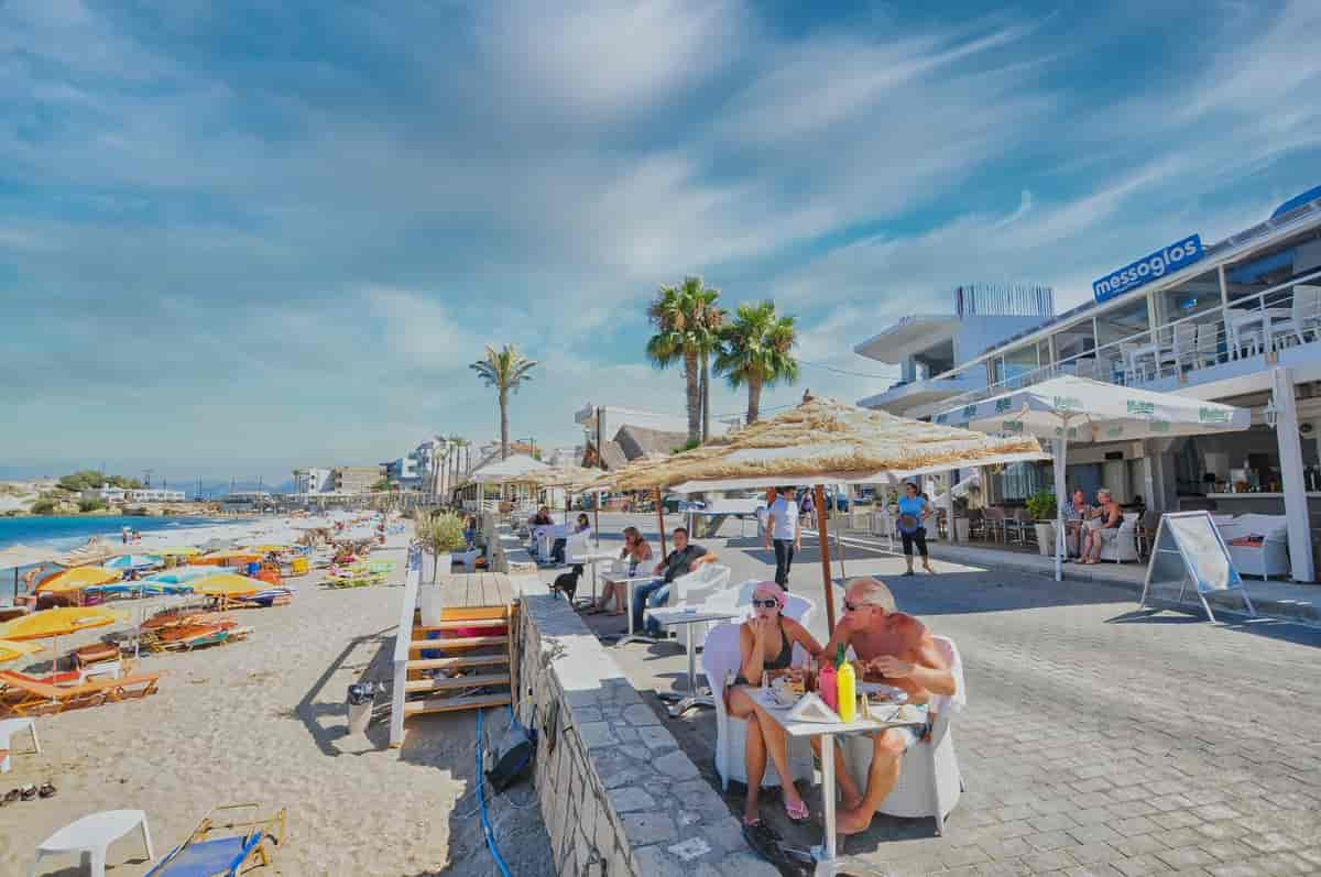 Turister sitter ved bord under parasoller ved en uterestaurant. Til venstre er det en strand med strandstoler. Foto