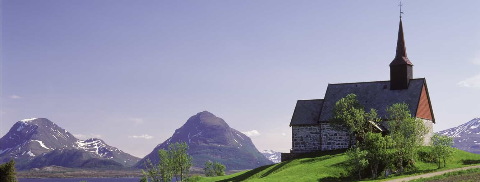 Fotografi av en liten kirke på en bakketopp. Kirken er av stein, og den har et spisst tårn av tre. I bakgrunnen ser man høye fjell. 