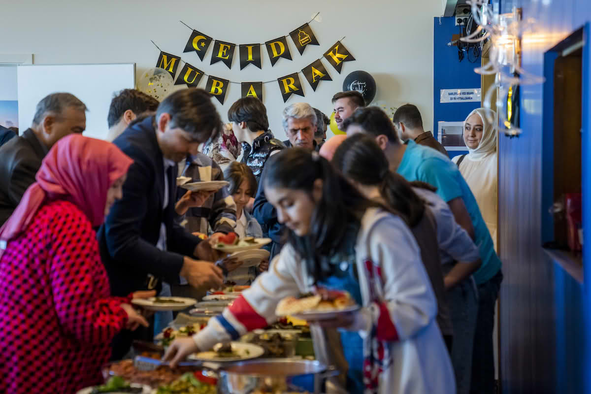 Flere mennesker samlet i et lokale. De står rundt et bord med mat og holder på å legge mat på tallerkner. På veggen bak henger det et banner hvor det står «eid mubarak».