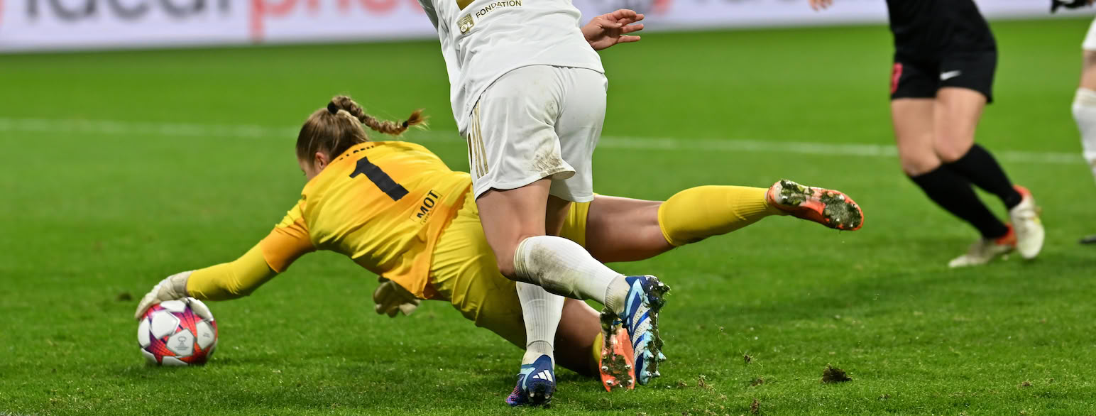 Foto av en keeper i gul fotballdrakt som slenger seg mot bakken som er dekket av grønt gress. Hun strekker venstre arm mot en fotball. Rett bak henne er en person i hvit fotballdrakt som står oppreist. Bare nederste del av kroppen er synlig. I øverste høyre hjørne er en person med svarte fotballstrømper og svart shorts.