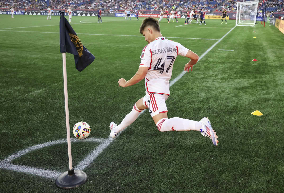 Foto fra hjørnet av en fotballbane inn mot mål og målområdet. I hjørnet står et flagg, og ballen ligger på en markert strek rett ved hjørnet. Spilleren som skal ta hjørnesparket er i fart og skal til å skyte med venstre bein. Foran målet står spillere på begge lag klare til å ta imot ballen.