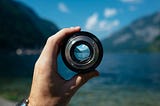 A hand grips a camera lens without the body of the camera attached. Through the lens you get a much more focused view of a lake and the mountains on the horizon.