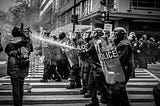 A wall of police in riot gear, head gear, and shields face a crowd and are spraying water onto the crowd.