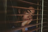 A young woman looks through some window slats. Her face is sad and cupped on her hand