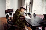 White woman wiht blond hair and a mauve hat writing on a notebook on a wooden desk and chair in a courtyard.