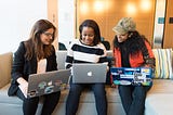 Three women sitting next to other, smiling, and looking at the middle woman’s laptop (all three have a laptop)