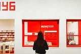 A woman stands in front of a bright red window inside a gallery shop.