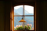 A view through a small window, divided into four by thin strips of wood, showing distant hazy blue hills and a mountain peak. Pink flowers in foreground outside the window. The walls around the window are dark, emphasizing the light outside.