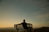 The back of a lady sitting on a bench. Her 2 arms are stretched on the backrest and she is looking at a big open field in front of her.