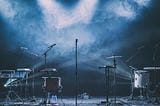 An empty stage, with drums, instruments and microphones under a haze of cloud. No one is on the stage.