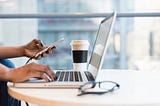 hand on a laptop with other hand holding phone and coffee cup in background