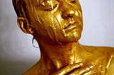 A photo of a person with short slicked back hair, covered in gold paint. It is dripping down their entire face and shoulders. Their eyes are closed and their skin is shiny gold