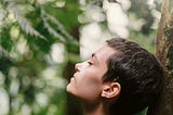 Lady with very short hair with eyes closed. Her head is slightly facing up and leaning against a tree.