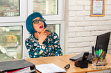 Office worker with blue hair thinking while sitting at a desk.