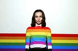 A woman stands in front of a rainbow backdrop with rainbows on her shirt.