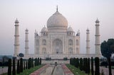 A large, square white marble structure with an onion dome topped by a finial. This dome is surrounded by four smaller domes. Four tall minarets stand at each of the four corners. The structure and the minarets all stand on a plinth. In the foreground is a long pool reflecting the structure with a row of trees on either side.