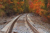 Railroad tracks turning around a bend lined by tall trees whose leaves have turned colors in Autumn