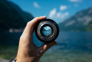 A hand grips a camera lens without the body of the camera attached. Through the lens you get a much more focused view of a lake and the mountains on the horizon.