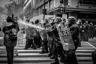 A wall of police in riot gear, head gear, and shields face a crowd and are spraying water onto the crowd.