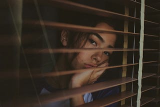 A young woman looks through some window slats. Her face is sad and cupped on her hand