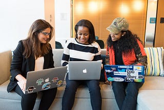 Three women sitting next to other, smiling, and looking at the middle woman’s laptop (all three have a laptop)