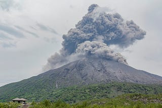 A volcanic eruption is depicted, with ash and smoke billowing from the summit against a backdrop of green hills and a cloudy sky.