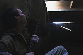 A man with a rifle takes cover behind a barricade.