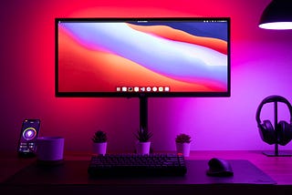 An LCD computer and peripherals on a desk. The computer displays a huge range of colors while the wall behind it is also lit in a reddish to purple spectrum — these colours often represent digital culture.