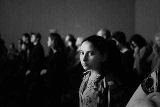 A girl looks at the camera. There is a quizzical expression on her face that could also be interpreted as blank. She is part of an audience that seems to be watching something.