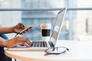 hand on a laptop with other hand holding phone and coffee cup in background