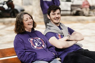 Two friends, both dressed in purple, sitting on a bench and laughing.