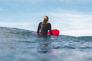 A female surfer clad in a wetsuit, sitting in the ocean on her red surfboard, waiting for waves