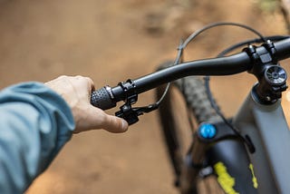 A photo of a man's left hand, reaching down to grasp the left handlebar of a bicycle. The handlebars are black, and we only see the left-hand side of the handlebars, with the front wheel of the bicycle out of focus in the background of the picture.