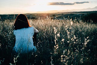 Female with short dark brown hair sitting in field in hilled area with sunset in the background.