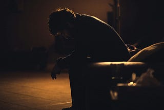 A young man sits alone in a darkened room, seemingly depressed and defeated.