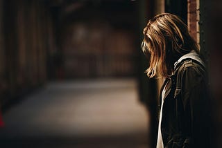 A vulnerable-looking young woman stands in an empty corridor, her head downcast.