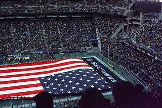 Image of a large American flag draped across a football stadium | Photo by Caleb Woods on Unsplash