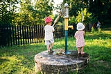 Two young children, wearing sun hats, stand by an outdoor water pump in a grassy area on a sunny day. One child is operating the pump handle while the other watches. The scene is peaceful, surrounded by greenery and a wooden fence in the background.