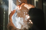 A person stands by a window facing the sunlight (though the room is dark) with her arms in the air as though they are catching sunbeams and welcoming them in.