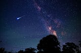 This is an image of the sky at night. There are a lot of bright stars in the sky. On the one hand, we can see the red moon. On the other hand, we can see an asteroid falling from space to the earth. The image has been taken from the ground where we can also see trees appearing black.