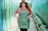 A woman with red long hair wearing a hat, green apron, and a smile.