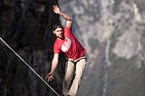 A casually dressed man in his thirties wears an intent expression as he catches his balance on a tightrope which is strung thousands of feet above rocky terrain.