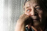 An elderly woman with crystal-clear blue eyes, sits next to a window but doesn’t look out of it.