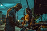 A bald man and a young woman stand next to a car with the hood open on a dimly lit street at dusk. The man, wearing a grey polo shirt, holds a wrench, while the woman, in a white tank top with her hair pulled back, watches closely. They appear focused on fixing the car together, with streetlights and power lines visible in the background, setting a moody evening scene.